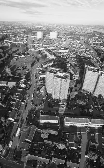 A black-and-white aerial photograph displaying a densely populated urban area with a mix of low-rise and high-rise residential buildings, streets, and parked vehicles. In the foreground, several multi